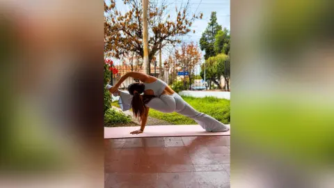 MirandaValentin poses in a deep stretch on a pink yoga mat, wearing a light blue sports bra and matching leggings outdoors.