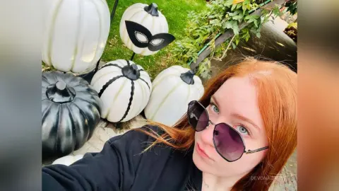 Redheaded woman in black outfit wearing sunglasses next to white and black decorative Halloween pumpkins.