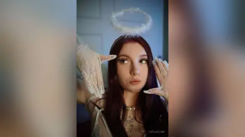 Young woman with dark hair wearing an angel halo and white lace gloves in a soft, ethereal indoor setting.