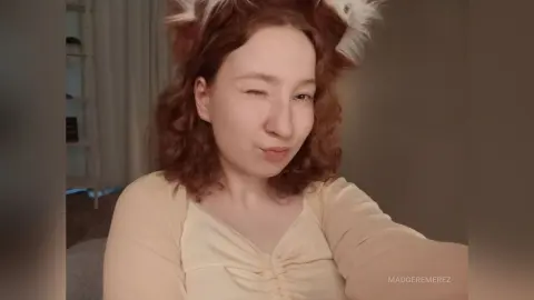 MadgeRemerez winks at the camera, wearing a light cream top and white furry headbands or ears, with her curly red hair down.