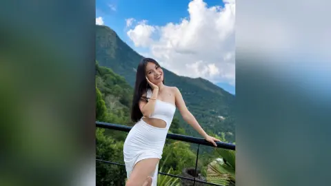 Smiling woman in a white summer dress posing against a scenic mountain landscape under a blue sky.