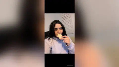 Young woman in a cozy lavender hoodie eating a snack in a bright indoor setting.