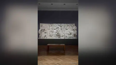 Large white classical relief sculpture depicting mythological figures mounted on a dark gallery wall.