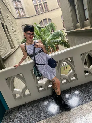 Stylish woman in a striped mini dress and glasses leaning on a stone balcony in a sunny courtyard.