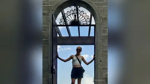 Blonde woman in a white tank top and shorts standing in a stone archway doorway under a bright blue sky.