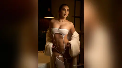 EvaCarusso poses in a white bra and thong, wearing an open white robe and white thigh-high stockings against a dark background.