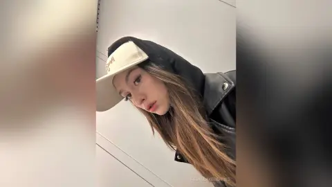 Young woman in a black leather jacket and cream baseball cap posing with a cool, edgy aesthetic.
