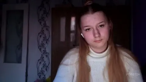 Young woman with long blonde hair wearing a white turtleneck sweater in a dimly lit room.
