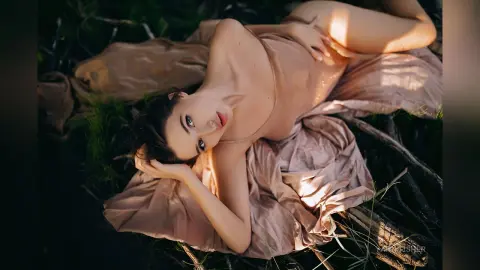 Elegant woman in a flowing beige silk dress lying in a dreamy, sunlit forest setting.