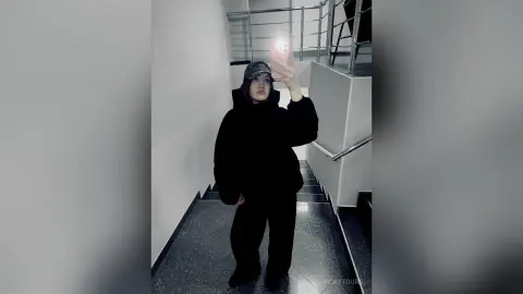 VickyToure poses in a black hoodie and black pants, wearing a dark baseball cap while taking a mirror selfie in a hallway with stairs.