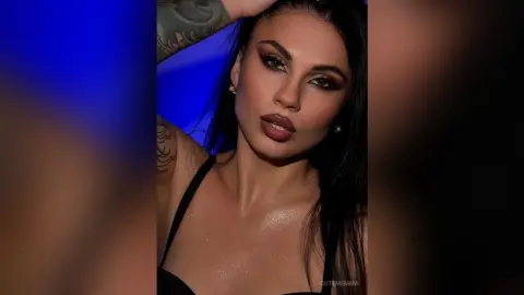 CutemEmma poses with long black hair and heavy eye makeup, wearing a black bra against a blue background.