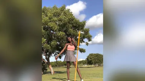 Stylish young woman in a pink tank top and grey mini skirt posing in a sunny green park under a blue sky.