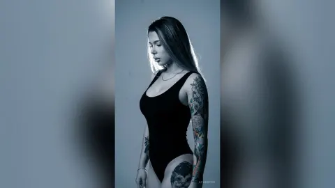 AvaBronX — AvaBronX poses in a black bodysuit, showing off her long hair and colorful arm tattoos against a plain background.