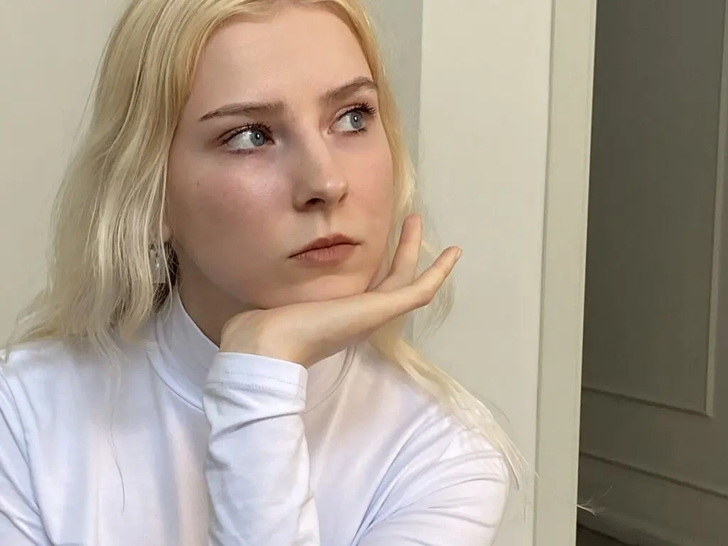 Blonde woman in a white turtleneck looking thoughtfully away in a bright indoor setting.