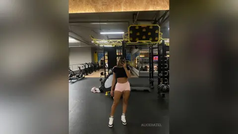 Fit woman in black crop top and pink shorts taking a mirror selfie in a modern gym setting.