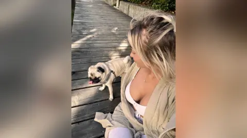Blonde woman in a beige cardigan sitting on a wooden boardwalk with a cute pug dog on a sunny day.