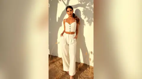 Stylish woman in a white linen crop top and wide-leg trousers posing in warm sunlight against a textured wall.