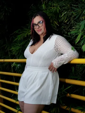 Young woman with glasses in a white lace romper posing against lush green bamboo foliage at night.