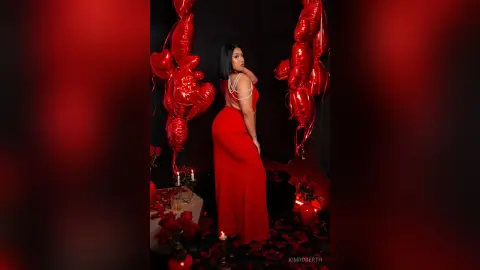 KimRoberth poses in a long red dress, standing against a dark background decorated with red heart-shaped balloons and rose petals.