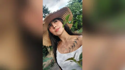RossiNoleen wears a large straw hat and a white tank top while looking up against a background of green trees.