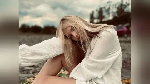 Blonde woman in a white linen shirt sitting in a dreamy outdoor field under a cloudy sky.