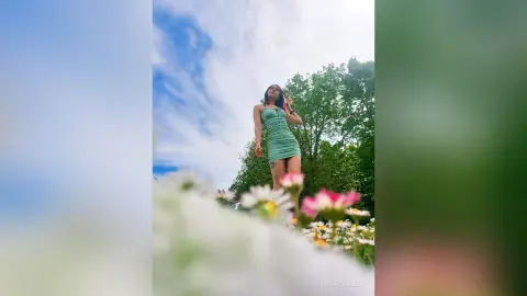 RossiNoleen stands outdoors in a tight green mini dress against a blue sky and green trees.