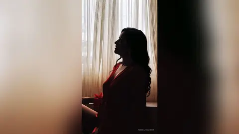 Silhouette of a woman in a red dress sitting by a window in soft, moody natural light.