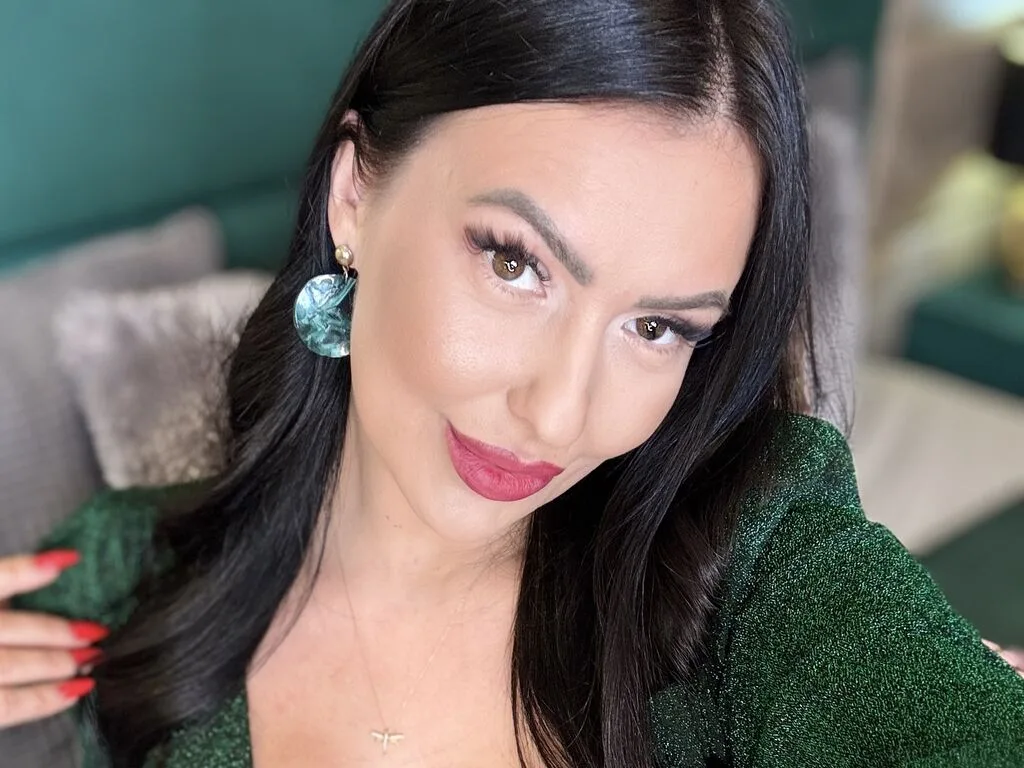 AlisonHale poses with long black hair and red lipstick, wearing a sparkly green top and large blue earrings.
