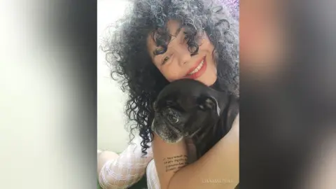 Smiling woman with curly hair hugging a small black dog in a bright indoor setting.