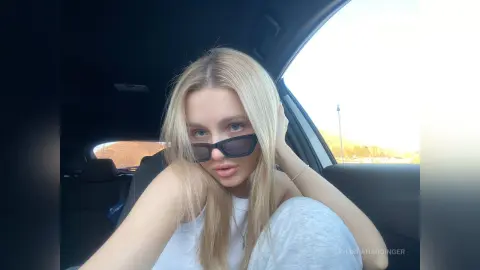 Blonde woman in a white tank top relaxing in a car during a sunny outdoor drive.