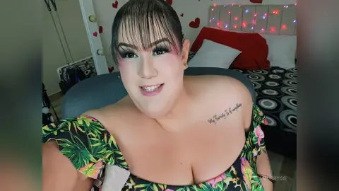Smiling woman in a tropical floral print dress with glam makeup in a cozy bedroom setting.