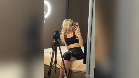 Blonde woman in a black crop top and leather skirt taking a mirror selfie in a cozy bedroom setting.