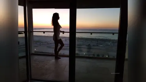 Silhouette of a woman in a white dress standing on a balcony overlooking the ocean at sunset.