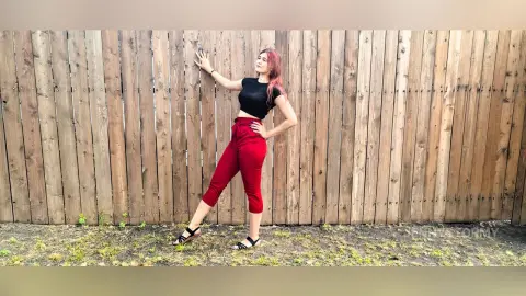 Redheaded woman in a black t-shirt and red trousers posing confidently against a rustic wooden fence.