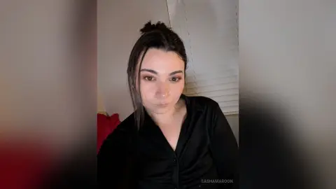 SashaMaroon looks toward the camera wearing a black button-up shirt with her dark hair pulled back in a messy bun.