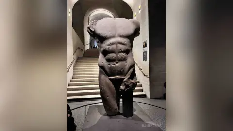 Ancient Greek marble torso sculpture displayed in a minimalist museum hallway with dramatic lighting.
