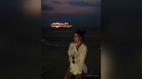 AnaMathers poses on a dark beach at night wearing a white button-down shirt and matching white shorts.