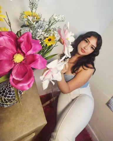 Young woman in white trousers sitting by bright pink and yellow flowers in a bright indoor setting.