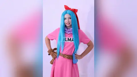 Smiling cosplayer in blue wig and pink shirt outfit posing against a light background.