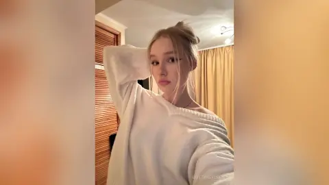 Blonde woman in a cozy white oversized sweater posing in a warm, sunlit room.