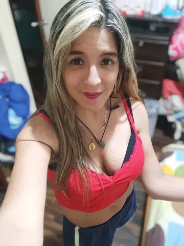 Smiling woman with blonde hair wearing a red crop top and gold necklace in a cozy bedroom setting.