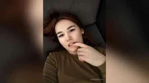 JaneBarbrick lies on a dark pillow with her hand near her mouth, looking into the camera with her brown hair spread out.