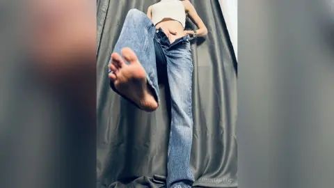 Young woman in white crop top and blue jeans relaxing against grey curtains in a casual indoor setting.