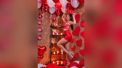 JessieLiu poses in a red lace bodysuit and red high heels, standing against a wall decorated with red hearts and white balloons.