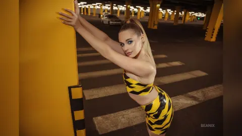 BeaNix leans back against a yellow pillar wearing a yellow and black striped tube top and matching skirt.