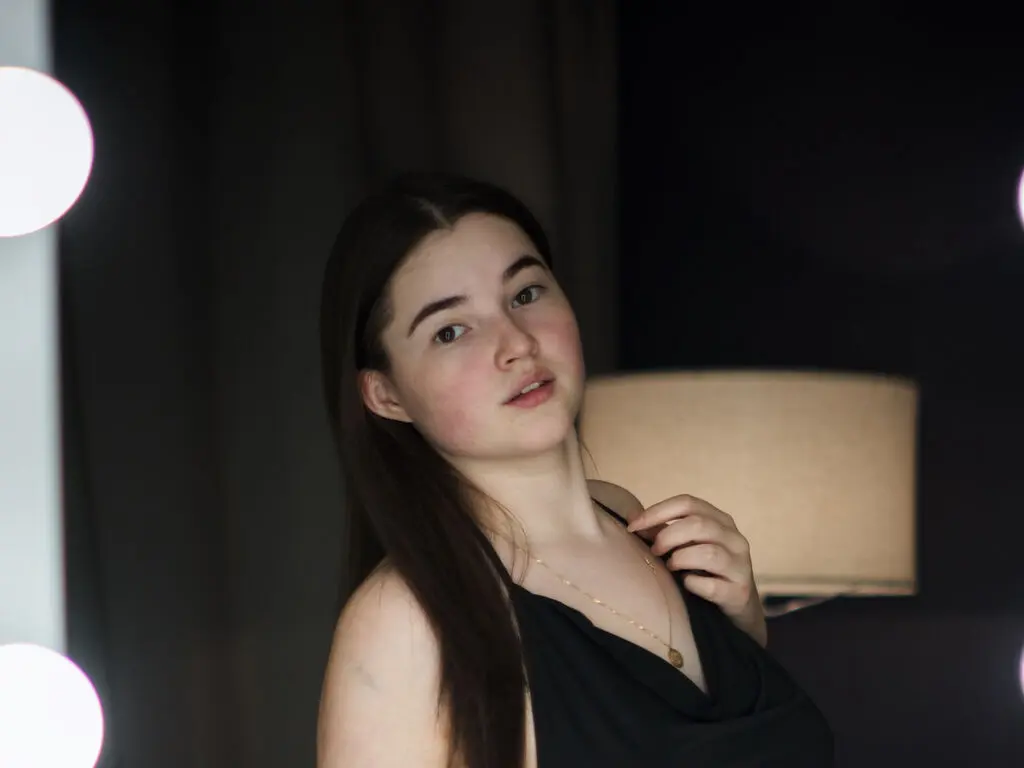 Young woman in a black tank top posing in a dimly lit room with warm ambient lighting.