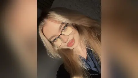 Smiling blonde woman with glasses wearing a navy blue shirt in warm, soft indoor lighting.