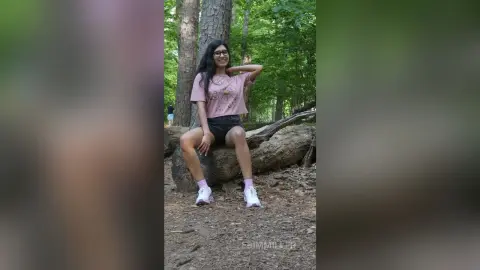 Smiling woman in pink graphic tee and black shorts sitting on a log in a lush green forest.