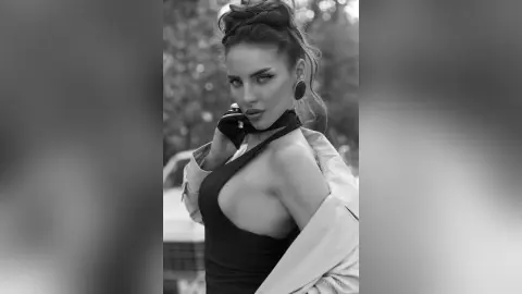 AnjeNoir poses outdoors in a low-cut black dress and an open white jacket, looking at the camera with her hair in an updo.