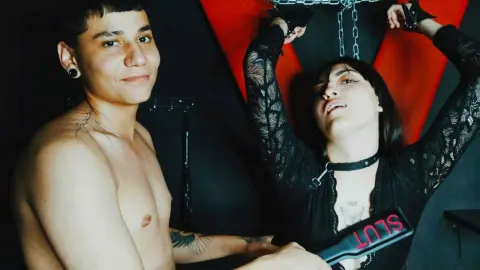 Shirtless man and woman in black lace lingerie posing with chains in a moody, dark studio setting.